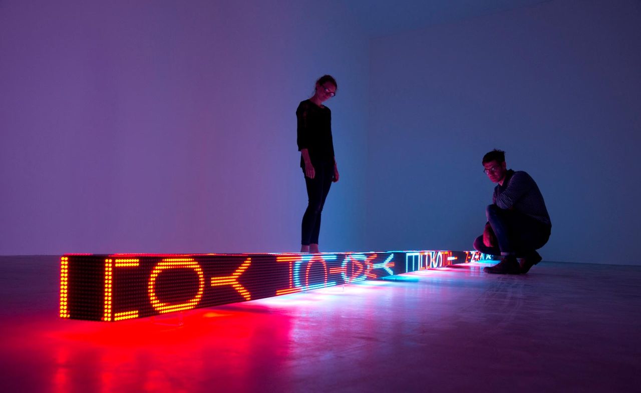 A man and a woman are standing next to the neon art installation, that&#039;s in a shape of a long beam with electronic neon writing on it.