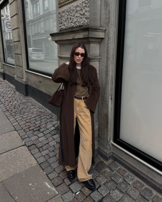 Annabel wears a brown coat, brown sweater, white t-shirt, tan pants, black boots, a brown bag and brown leather belt.