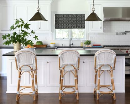 Bright, white kitchen with large island with black granite countertop and white based with cupboards, three bar chairs in rattan, two black cone pendant lights hanging over island