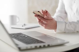 A person sat at a desk, with a laptop, using a smartphone