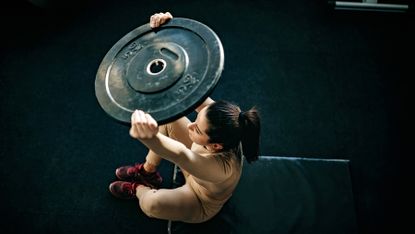 Woman lifts a weight plate while sat on a gym bench