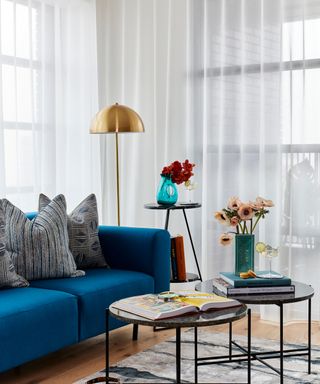 Modern apartment lounge with blue sofa, round nested tables, brass floor lamp, and sheer drapes.