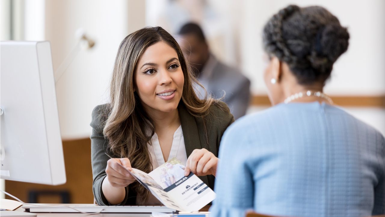 A woman behind a desk shows a credit union pamphlet to another woman.