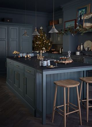 dark blue kitchen with kitchen island, wood bar stools, Christmas decor, Christmas tree on the countertop, garland across the range mantel