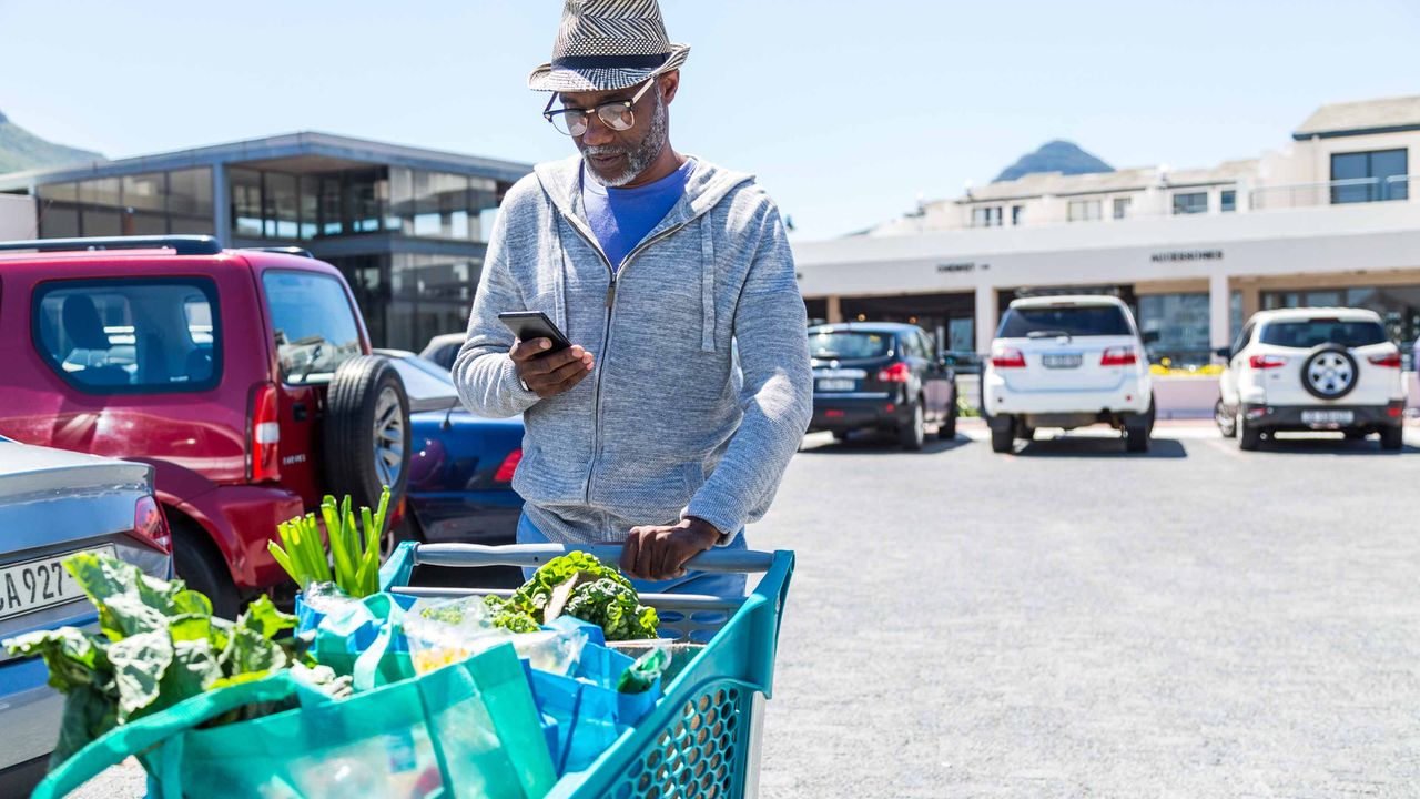 Retiree pushing full shopping cart while looking at smart phone