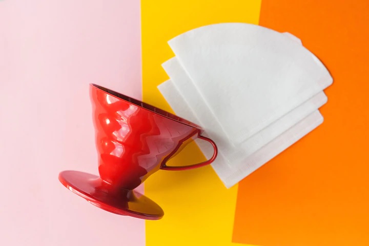 Un goutteur de café Hario V60 rouge, placé à côté de filtres blanchis sur un fond coloré