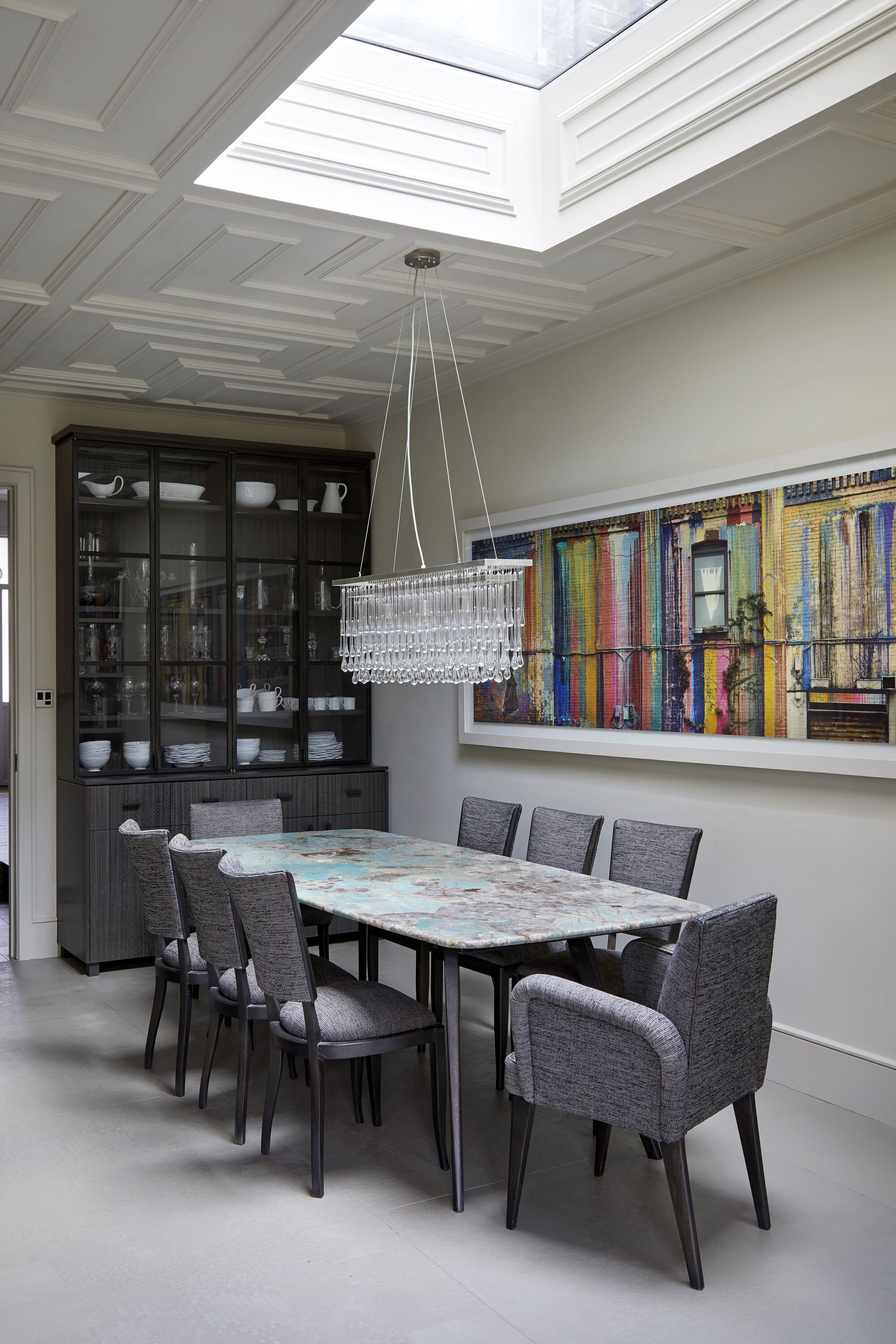 modern dining room with stone rectangular dining table surrounded by eight upholstered gray dining chairs, gray tiled floors, glass chandelier over dining table, hutch with china in it, colorful artwork, and decorative ceiling with large skylight