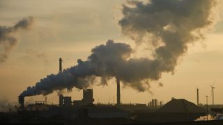 A smokestack billows thick gray smoke against a smoggy yellowish sky with the buildings below darkened in the haze