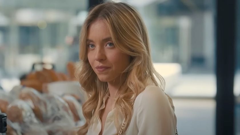 Sydney Sweeney standing in a coffee shop almost smiling in Anyone But You.