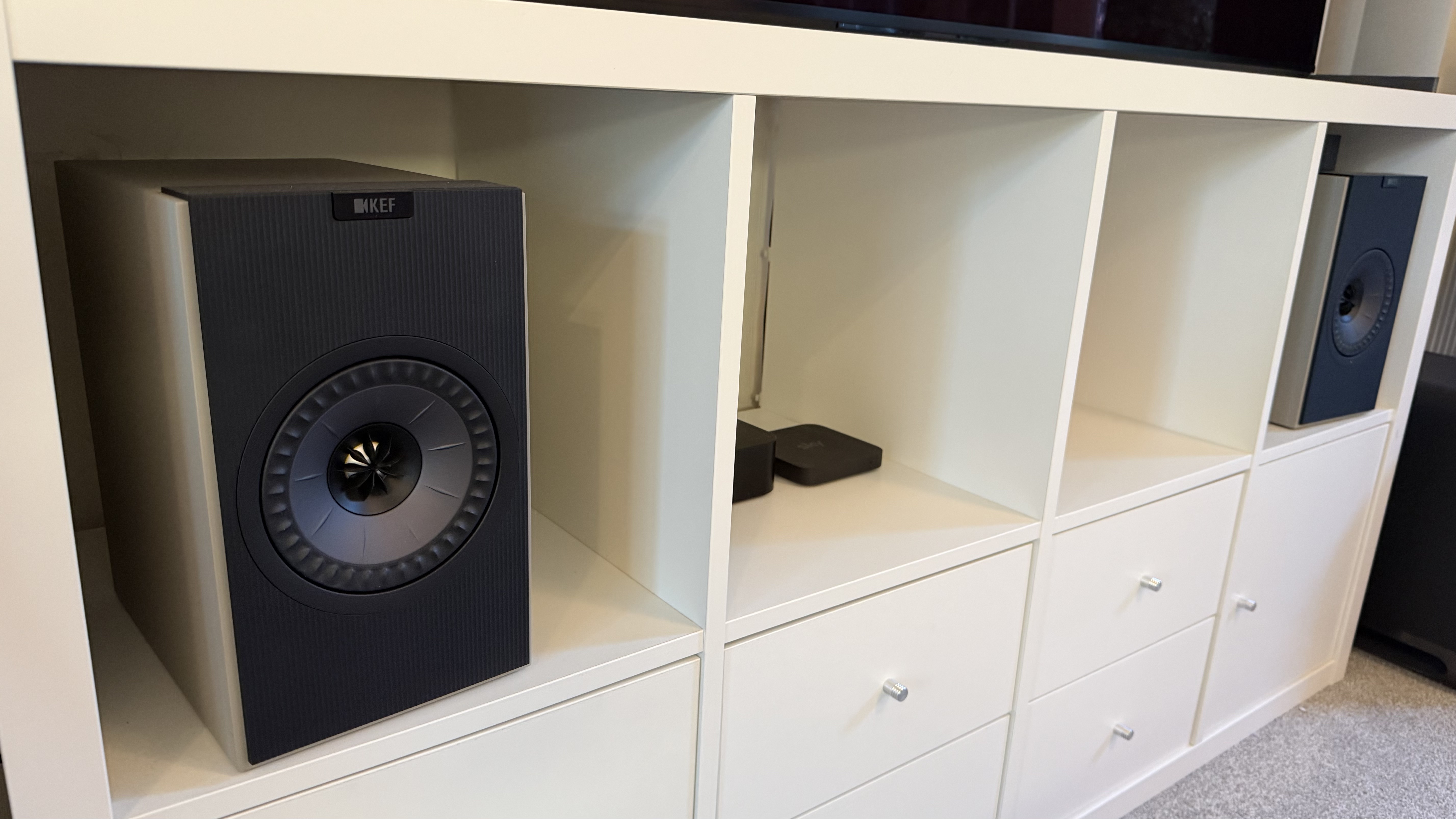 A white bookcase, upon which is standing an OLED TV, and in the nooks of which are a pair of KEF Coda W speakers, a Nintendo Switch, and a couple of other smaller components