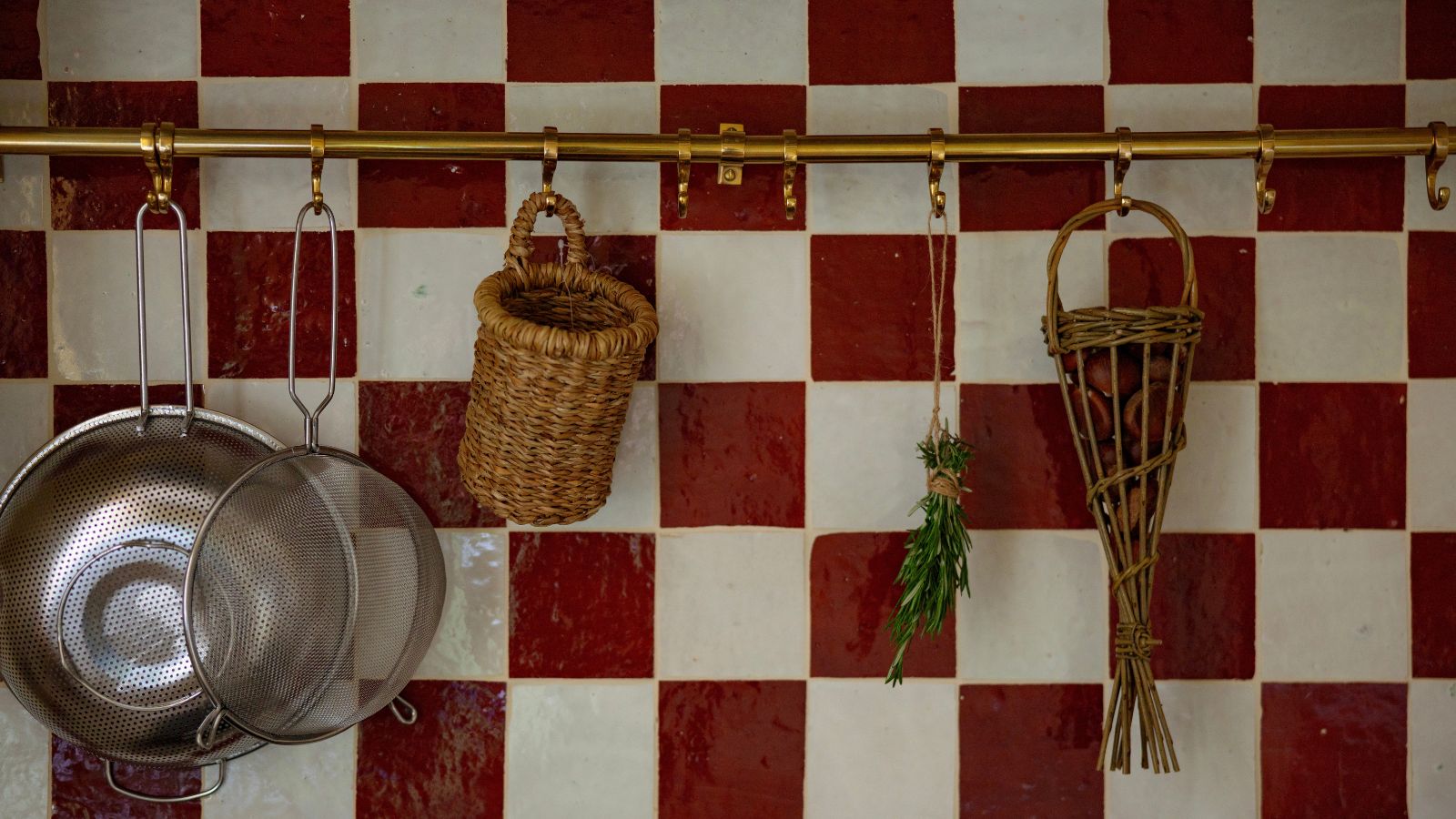 Handmade red and white zellige checkerboard tiles with brass utensil rail, colanders, and woven baskets in a coastal Shaker kitchen.