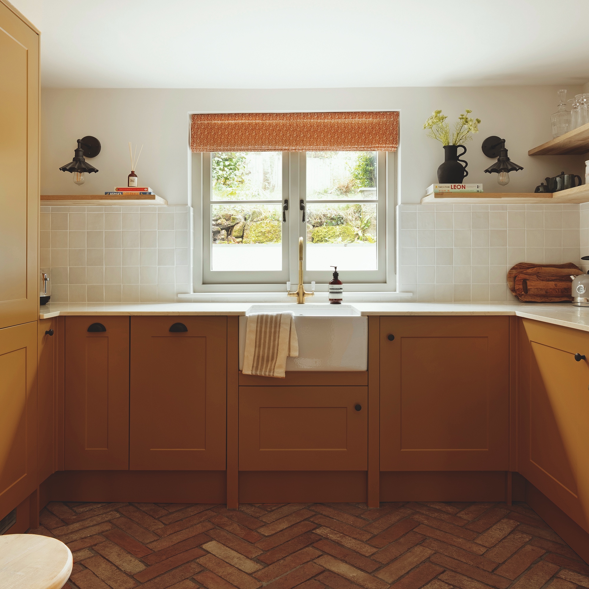 kitchen with shaker style deep yellow painted units, butler sink, square window with fabric blind and herringbone brick flooring