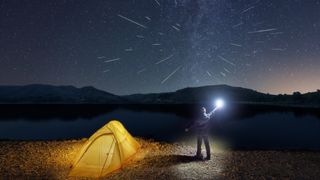A person stands next to a small yellow tent and holds up a bright light to the sky. Meteors streak across the sky in the background. 