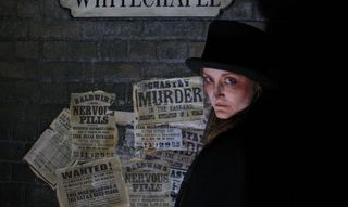 woman in dark clothes next to a wall, with 'murderer' written on papers behind her