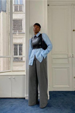 How to make a button-down shirt look more expensive is shown in a photo of a woman standing inside a home wearing a light blue button-down shirt under a black leather corset top styled with gray wide-leg trousers, black heels, and a black shoulder bag