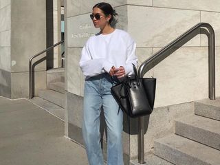 Influencer Sasha Mei poses outside a city building wearing aviator sunglasses, a long-sleeve white sweatshirt, baggy straight-leg jeans, and a black top-handle bag.