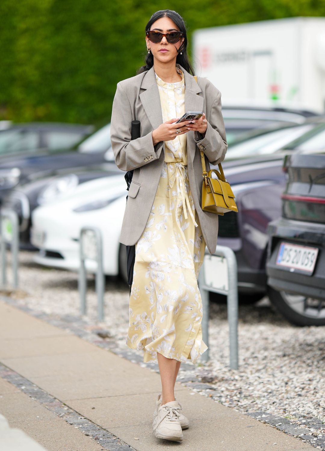 A guest wears black sunglasses, a pale gray long blazer jacket, a pale yellow with white embossed pattern long dress, a yellow shiny leather long handbag from Jacquemus, beige matte sneakers , outside Munthe, during the Copenhagen Fashion Week Spring/Summer 2024 on August 10, 2023 in Copenhagen, Denmark.