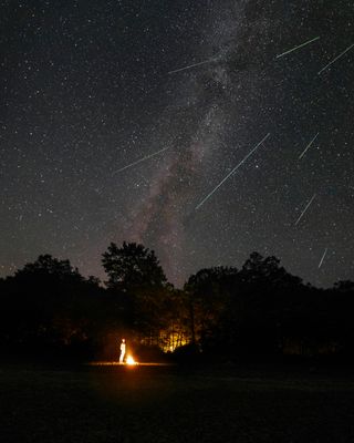 A humanoid figure stands next to a campfire amidst trees. He gazes up at the starry night sky, where streaks of shooting stars stretch across the tall milky way.
