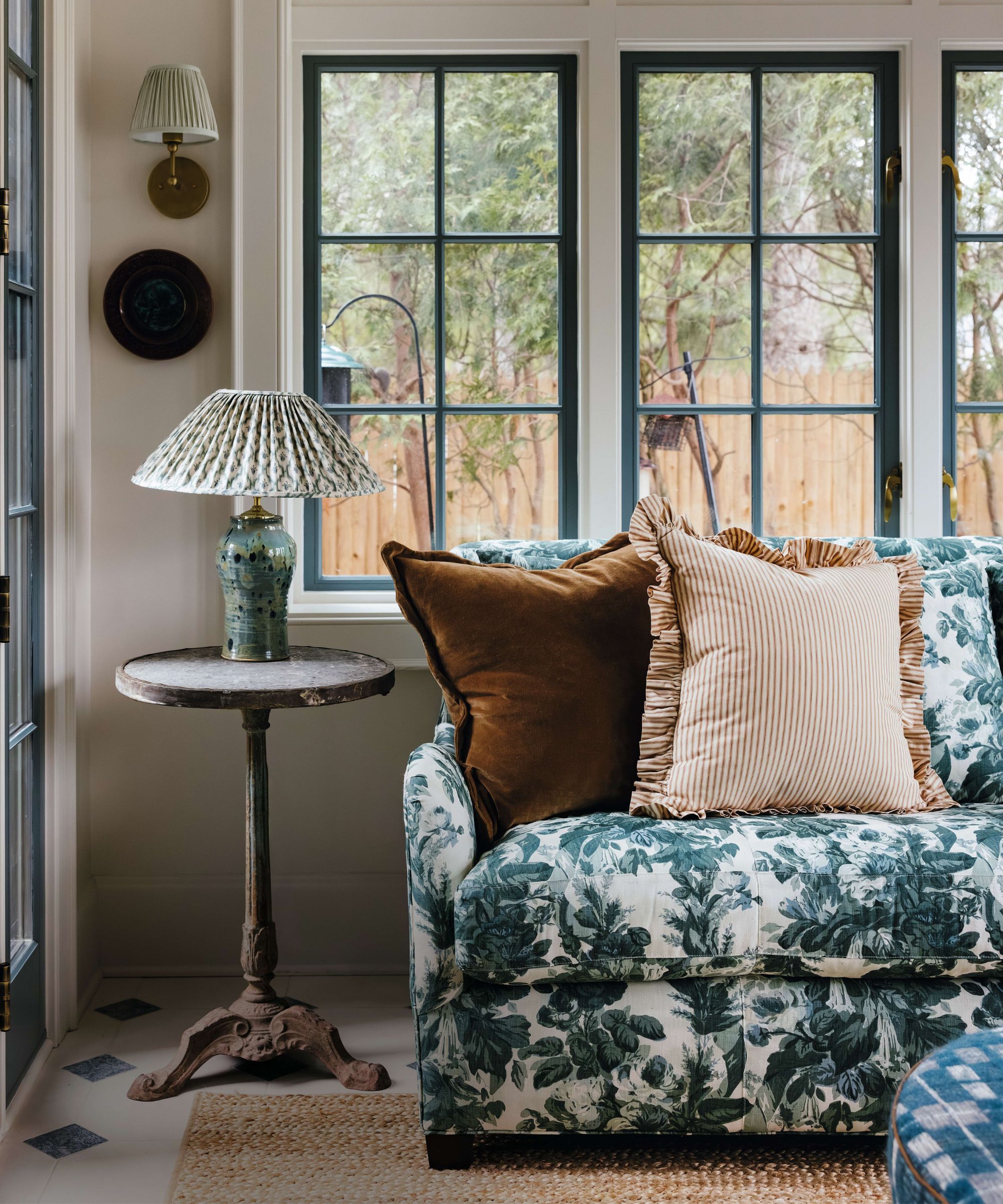 a neutral sunroom with floral chintz sofa and gree windows, woven rug, antique table and lamp