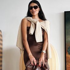 influencer and entrepreneur Babba Rivera poses in oversize oval sunglasses, a brown slip dress, a beige cable knit sweater around the shoulders, and a brown clutch bag