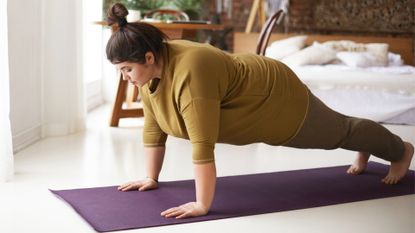 woman wearing khaki top and leggings in a bedroom setting performing a high plank on a purple mat.