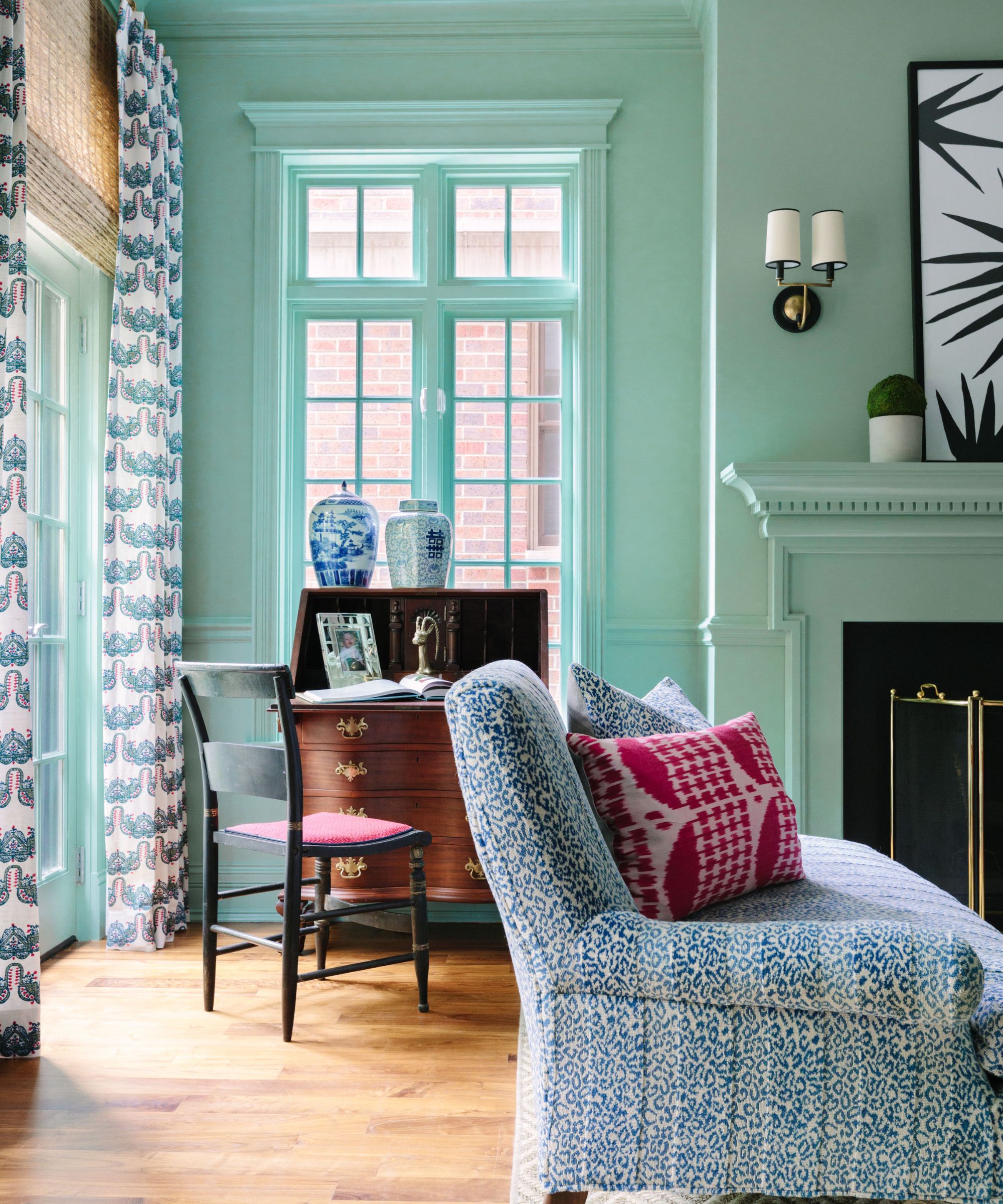 Living room painted mint green, an antique desk with a cane chair, a patterned blue sofa, glass doors with floral blue drapes, and a large fireplace.