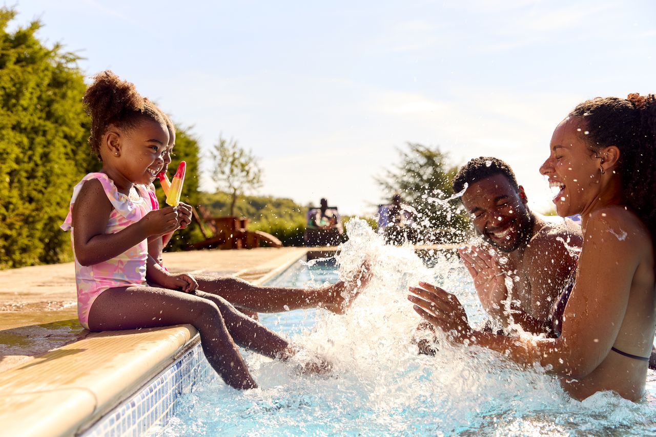 Family on summer holiday with two girls eating ice lollies by swimming pool splashing parents