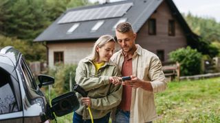 male and female stood in front of property with solar panels, plugging in EV charger into car while looking at smartphone