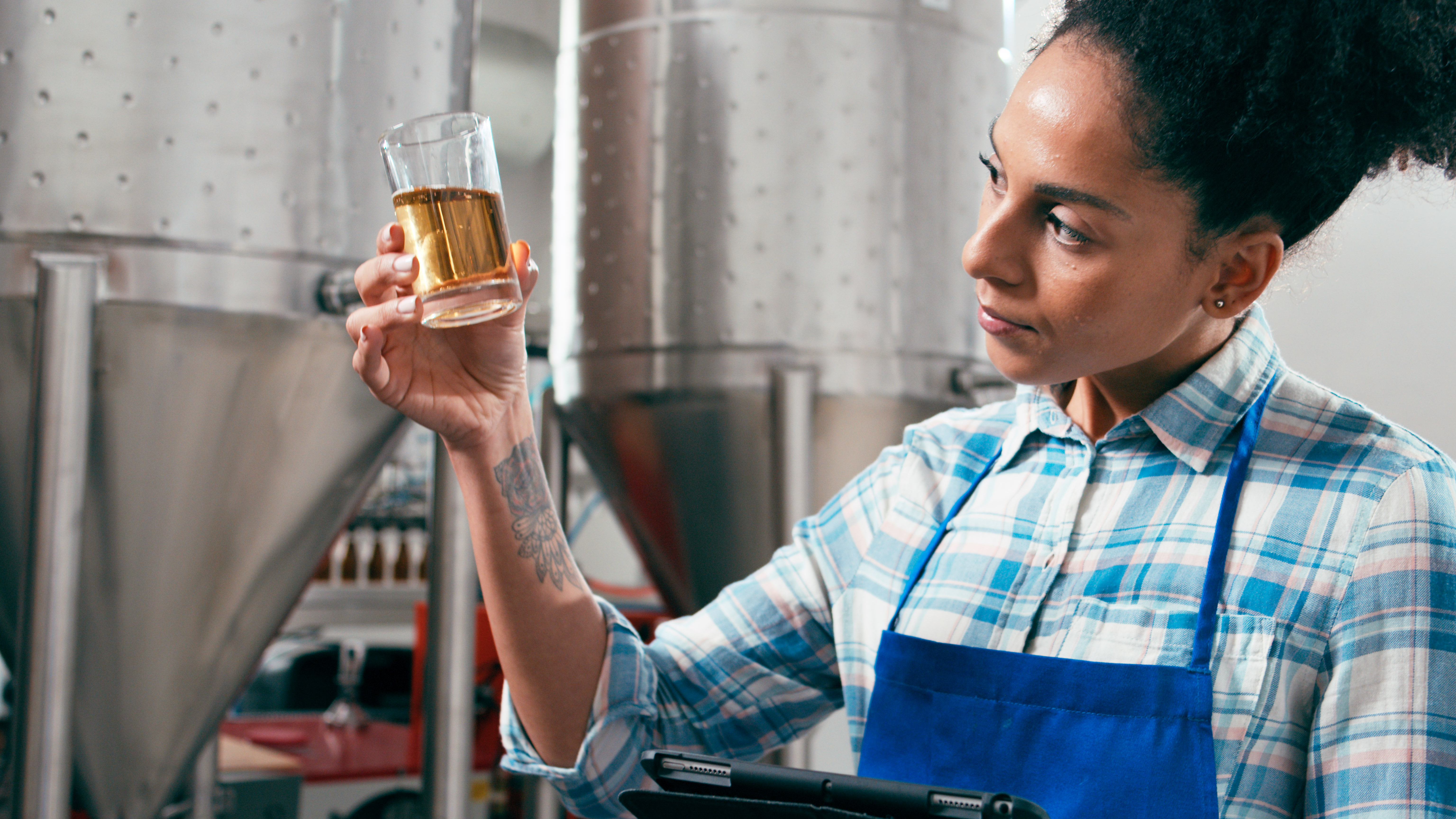Woman in checked shirt and apron inspecting a beer glass in a brewery