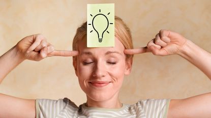 A smiling woman has a drawing of a lightbulb on a Post It Note stuck to her forehead.
