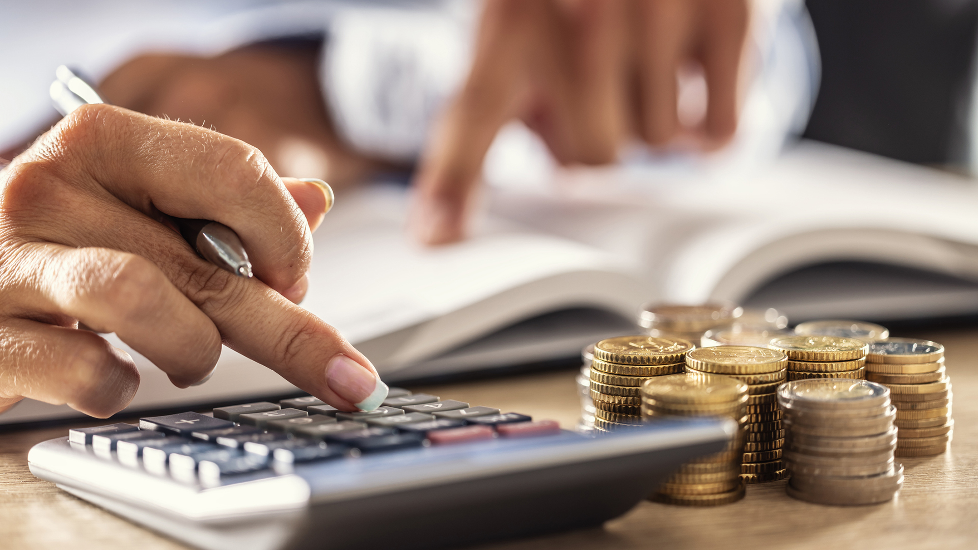 A close-up of a calculator being used by a business professional, with coins to the side of the calculator and a sales book open in the background.