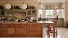 A bright kitchen with marble countertops, wooden and sage green cabinetry, large windows, striped blinds, white pendant lamps, flowers, and light tiles.