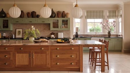 A bright kitchen with marble countertops, wooden and sage green cabinetry, large windows, striped blinds, white pendant lamps, flowers, and light tiles.