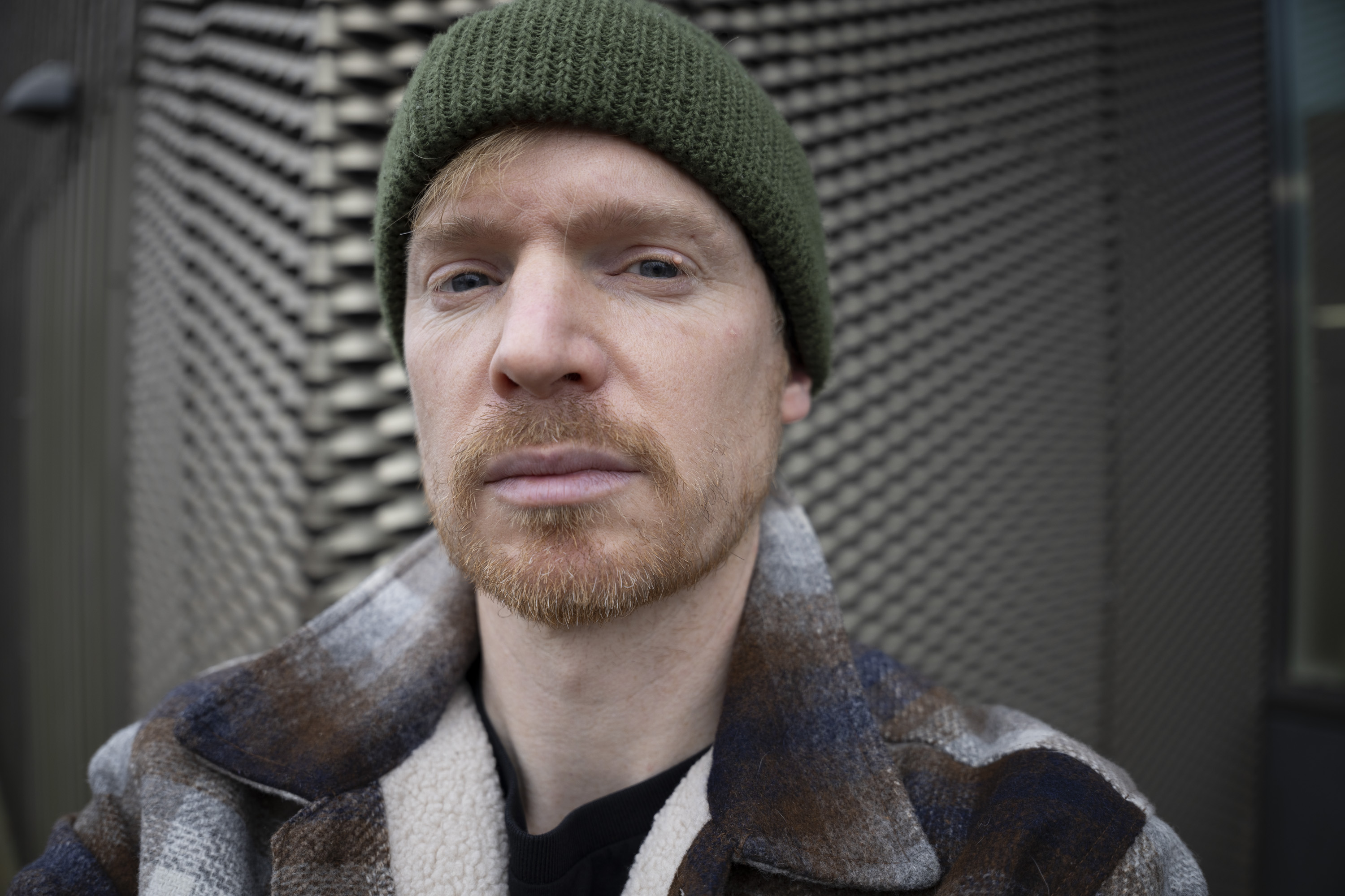 A man in a green beanie and shacket's selfie, outdoors in gray urban area