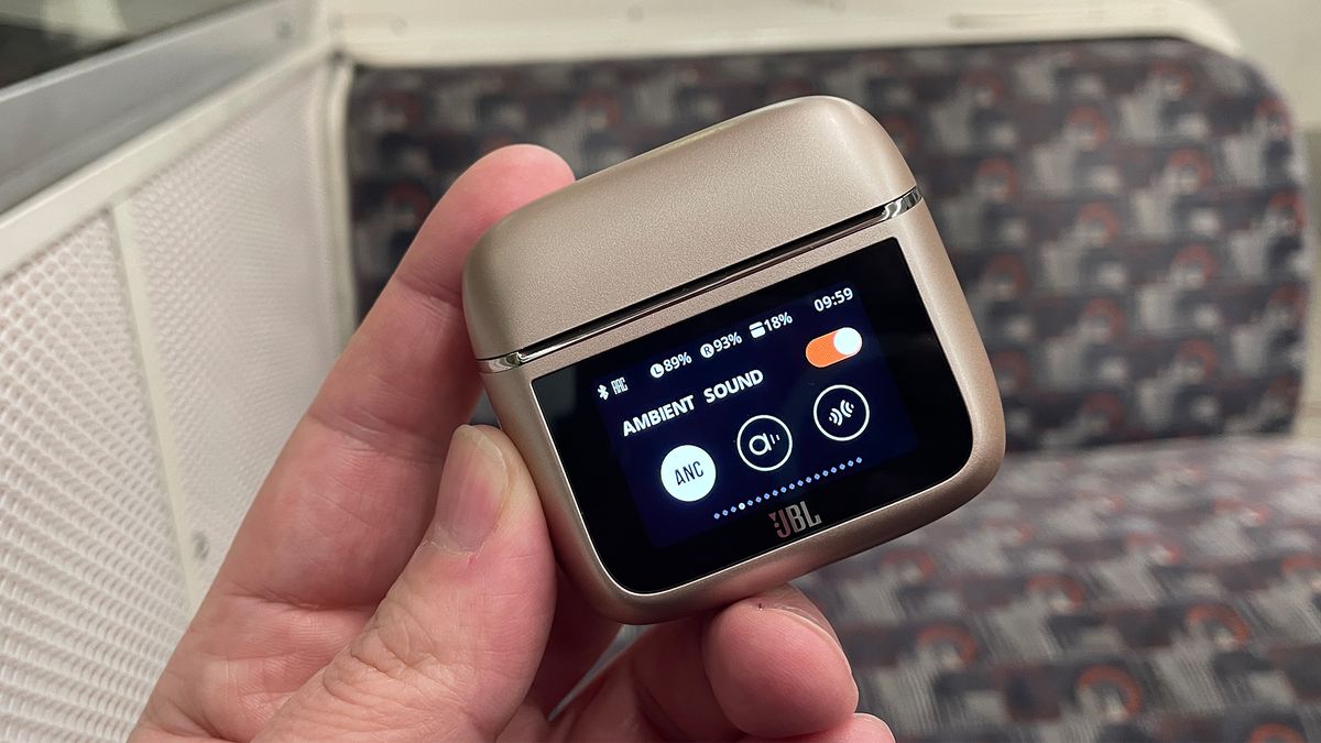 A hand holding the case for the JBL Tour Pro 3 earbuds. The lid is closed and the screen on the front is visible. It is showing the ANC settings options.