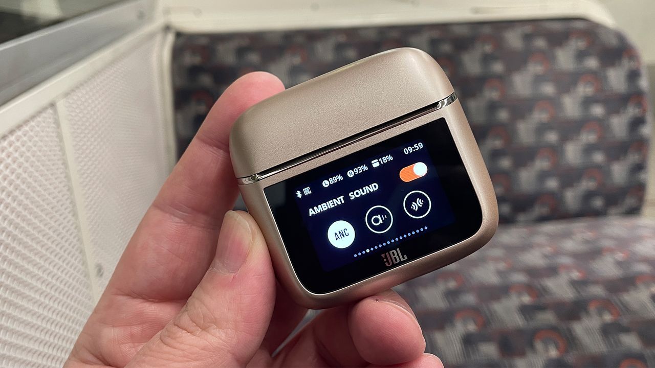 A hand holding the case for the JBL Tour Pro 3 earbuds. The lid is closed and the screen on the front is visible. It is showing the ANC settings options.