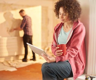 A lady sits in the foreground of a renovation project with an iPad and a cup of tea. She is sitting next to paint strappers and wearing a pink hoodie and blue trousers, both marked with paint, suggesting she has been renovating her home. A man stands in the background, stripping wallpaper from the walls.