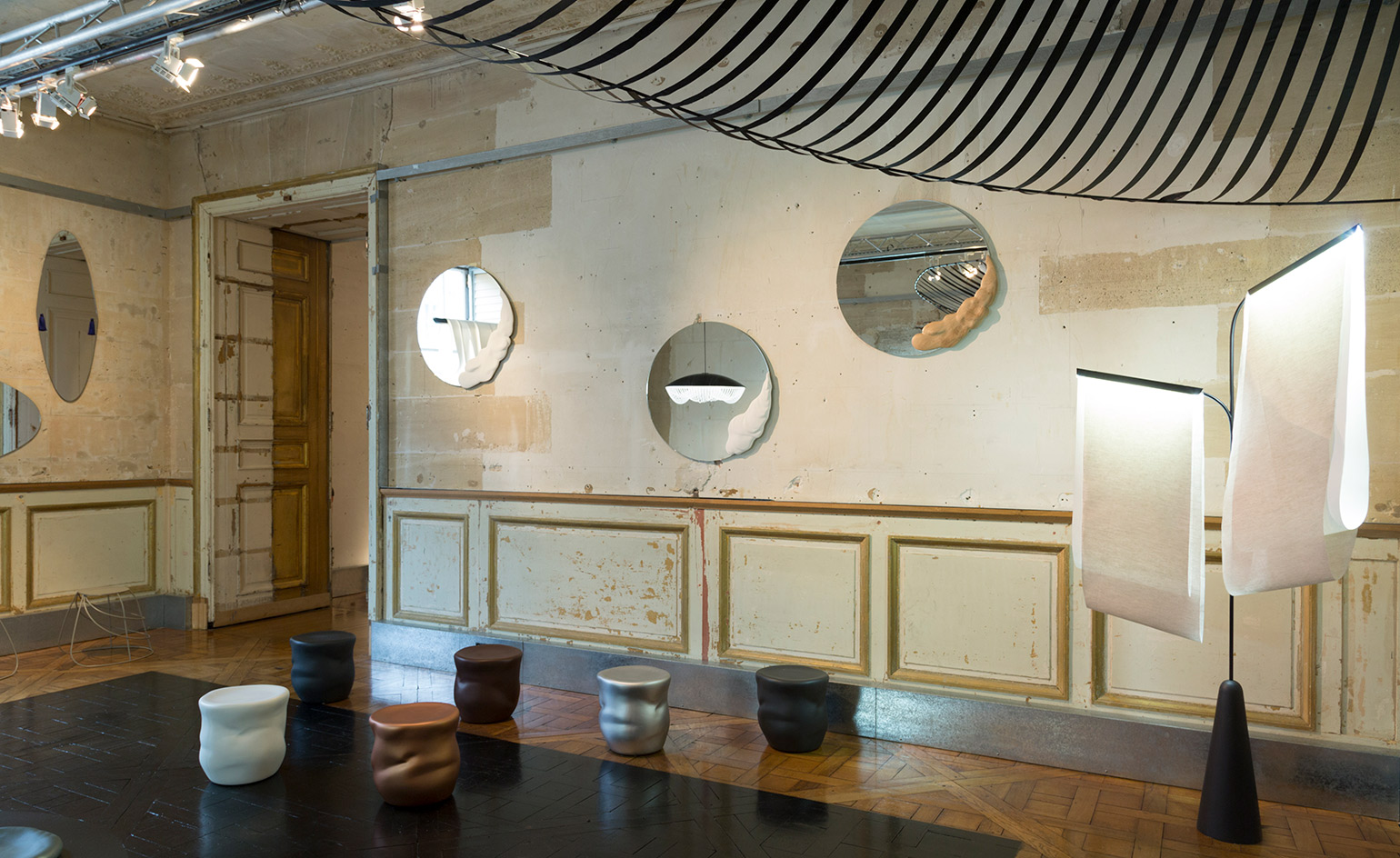 A room with run-down walls, with mirrors of various shapes set on each wall. Stool-like sculptures in different colors are set throughout the room.