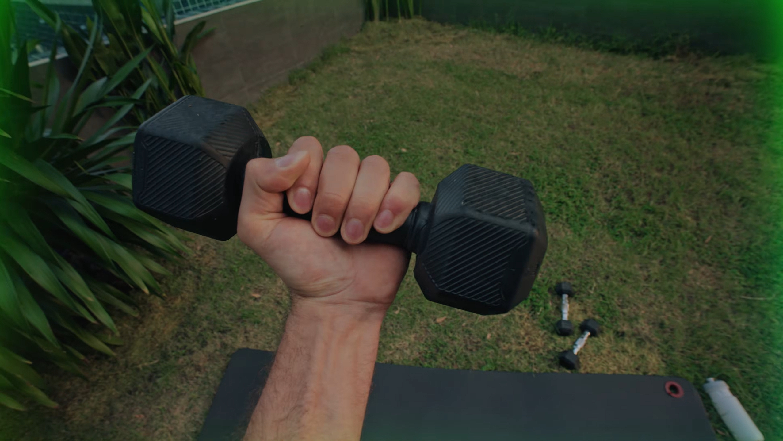 A close-up of a hand gripping a black hexagonal dumbbell against a grassy background and a dark yoga mat.