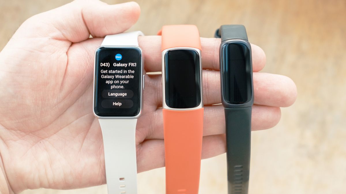 Close-up of the Samsung Galaxy Fit3 fitness tracker in hand next to a Fitbit Charge 6 with an orange strap and a Fitbit Inspire 3 with a black strap