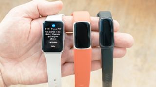 Close-up of the Samsung Galaxy Fit3 fitness tracker in hand next to a Fitbit Charge 6 with an orange strap and a Fitbit Inspire 3 with a black strap