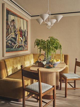 Image of a bright, beige breakfast room. There is a mustard yellow, velvet corner booth seat with a light wood table in front of it. There are two wooden dining chairs with blue cushions on the other side of the table.