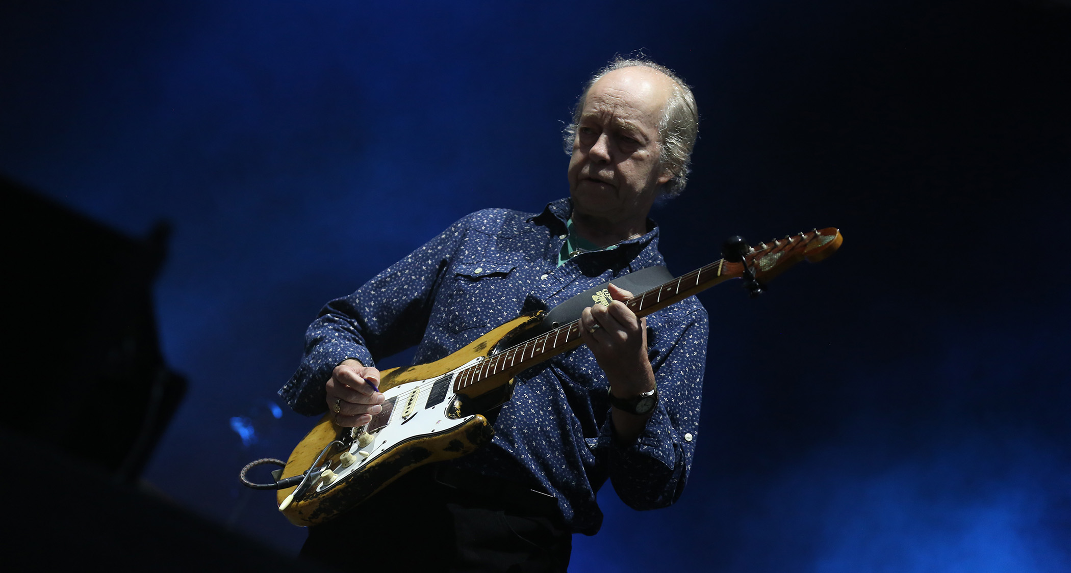Eric Bell wears a blue patterned shirt and plays his trusty Fender Stratocaster live in 2019