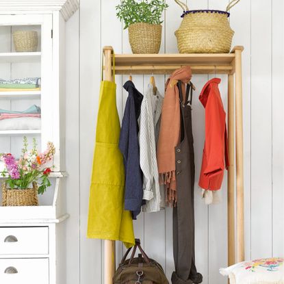 Wooden clothes rail hung with clothes against a white wooden wall beside a white dresser
