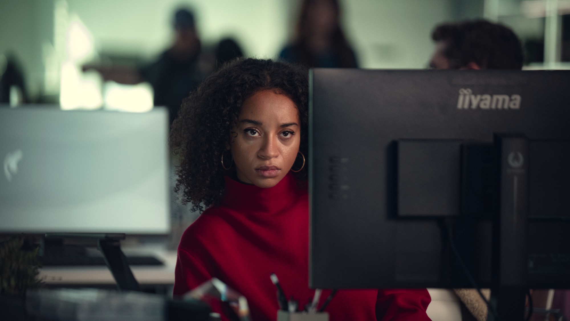 The actress Siena Kelly sits at a desk with her face looking at the camera / between two computer monitors. she is wearing a red turtleneck