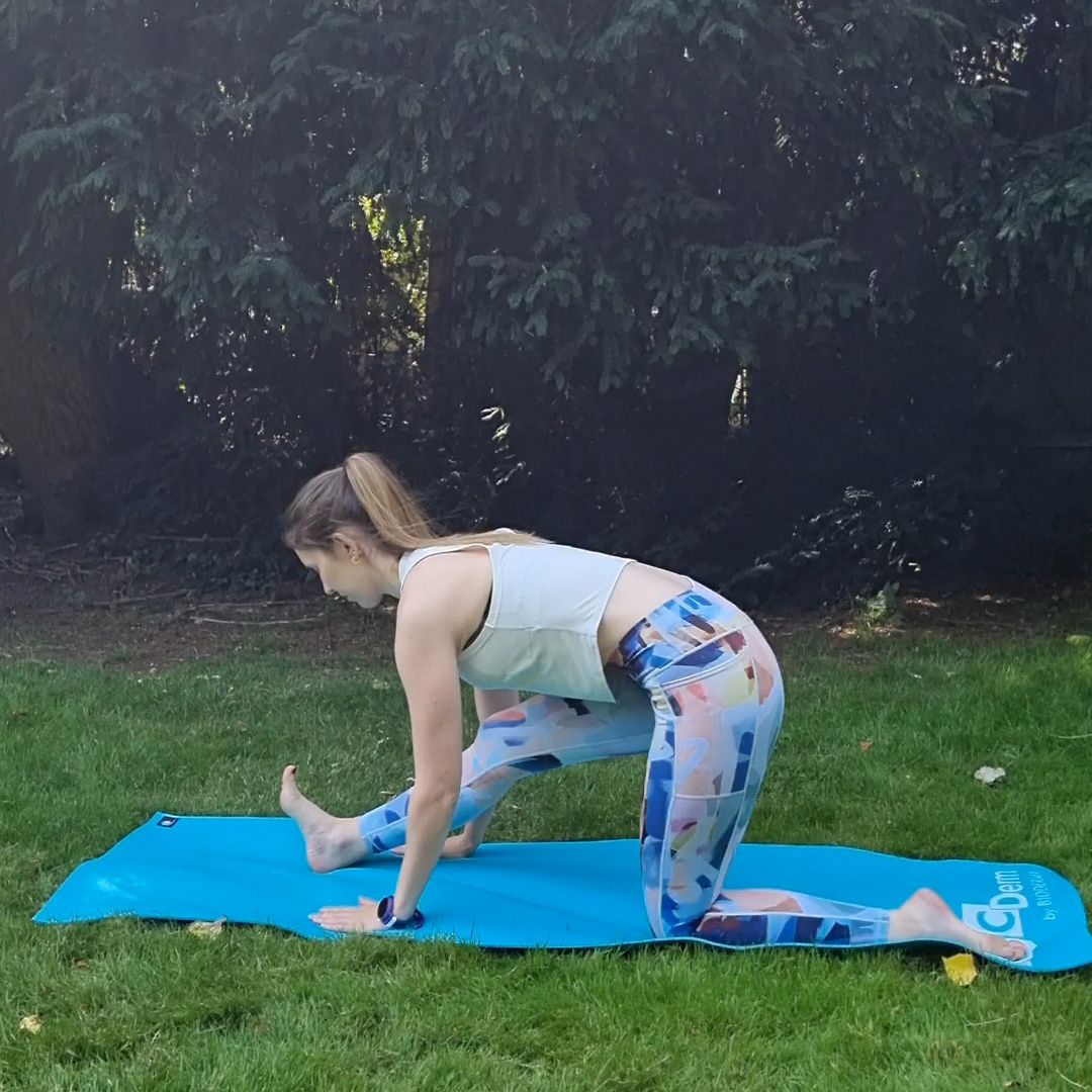 Fitness editor Ruth Gaukrodger performs a low lunge to half split. She is in sportsgear and exercising outside on a yoga mat. In this picture, she performs a half-split. Her right heel is on the ground and her right leg straight, while her left leg is bent at a right angle and her right shin running along the ground. Her torso is bent over her right leg and her hands are planted either side of her forward foot.