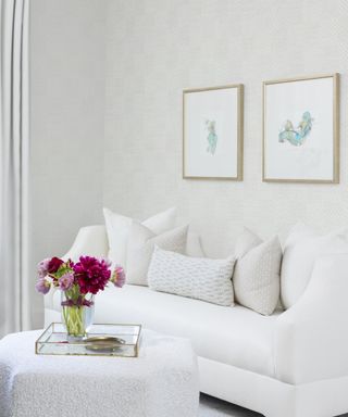 Room with white sofa and assorted throw pillows, upholstered ottoman and pale patterned wallpaper