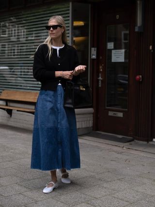 Eyewear designer and fashion influecner Linda Tol wearing a black cardigan over a white tee, a long A-line denim skirt, white Mary Jane–style ballet flats, and sunglasses.