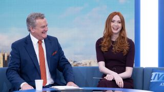 Hugh Bonneville as Douglas in a suit sits on a sofa next to Karen Gillan as Madeline in a maroon dress against a London backdrop in Douglas is Cancelled