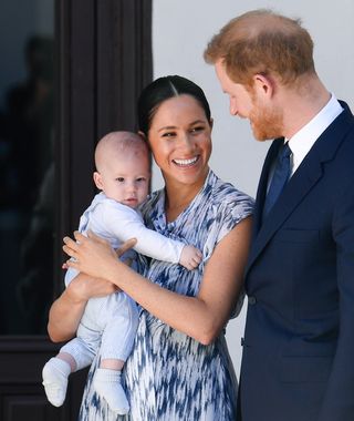 Prince Harry wearing a blue suit standing next to Meghan Markle in a blue patterned dress holding baby Prince Archie and smiling